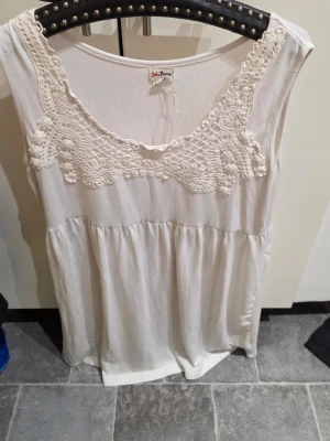 Vit ärmlös klänning med spets - Söt vit klänning från John Baner med ärmlös design och vacker spetsdetalj runt den djupa ringningen. Tillverkad i mjukt bomullsmaterial, perfekt för varma dagar. Klänningen har en avslappnad passform och lätt rynkad midja för en luftig känsla.