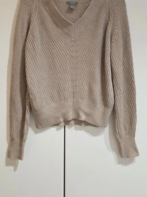 Beige stickad tröja från Lindex - Säljer en beige stickad tröja från Lindex med v-ringad hals och bred ribbad mudd vid ärmslut och nederkant. Tröjan har en avslappnad passform och är gjord i ett grovt stickat material som känns både trendigt och bekvämt.