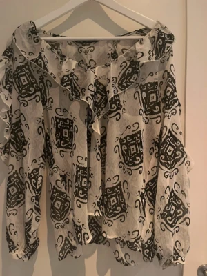 Mönstrad blus med volangkrage från Zara - Snygg blus i transparent vitt tyg med svart grafiskt mönster. Blusen har volangkrage och långa ärmar med resår vid ärmslut. Materialet är lätt och luftigt, perfekt för en trendig look. Passar dig som gillar unika detaljer och mönster.