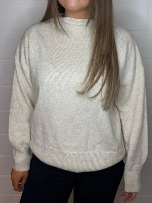 Beige stickad tröja med hög hals - Mysig beige stickad tröja med hög hals och avslappnad passform. Tröjan har långa ärmar och ribbade muddar vid ärmslut och nederkant. Perfekt för lager-på-lager och enkel att matcha med olika outfits.