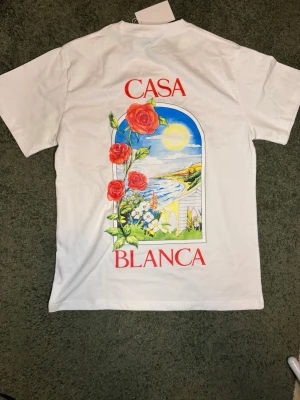Vit Casablanca t-shirt med rosor - Vit t-shirt från Casablanca med färgglatt tryck av rosor och landskap på både fram- och baksida. T-shirten har rund hals, korta ärmar och en avslappnad passform. Perfekt för dig som gillar unika prints och stilrena plagg.