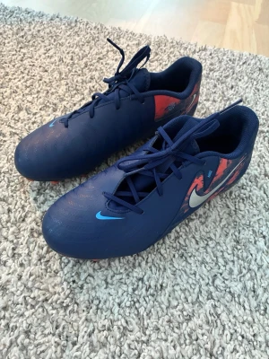 Nike Phantom GX fotbollsskor  - Säljer ett par blå fotbollsskor från Nike (Phantom GX). Junior. Skorna har snörning och är tillverkade i syntetmaterial med dubbar för bra grepp och kontroll på planen. Passar på gräs och konstgräs. De är använda några fåtal gånger. Benskydd på bild ingår.