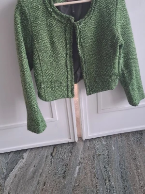 Grön stickad cardiganjacka - Grön stickad cardiganjacka med melerad struktur och rundad halsringning. Jackan har långa ärmar och en kortare, boxig passform. Materialet är grovstickat och ger en mysig känsla. Perfekt för lager på lager och att slänga över en t-shirt. Storleksmässigt som en medium.