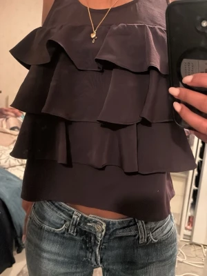 Mörklila volangtopp med ärmlös design - Snygg mörklila ärmlös topp med flera lager volanger framtill. Toppen har en loose passform och är perfekt att styla med jeans eller kjol. Materialet känns mjukt och har ett lätt glansigt utseende. Strl: S