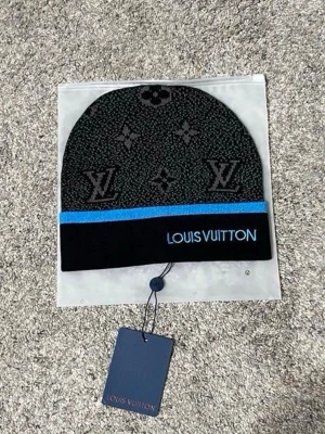 Svart mössa med blå text - Snygg svart mössa med ikoniskt monogrammönster och blå rand. Märkets logga är broderad framtill. Perfekt accessoar för dig som vill ha en exklusiv och trendig look. Materialet känns mjukt och bekvämt.