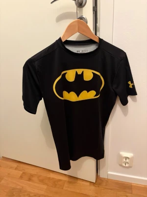 Svart Batman t-shirt från Under Armour - Cool svart t-shirt från Under Armour med stort gult Batman-tryck på bröstet. Modellen är tight och har korta ärmar, perfekt för dig som gillar superhjältar och sportig stil. Materialet är polyester och funkar grymt till träning.