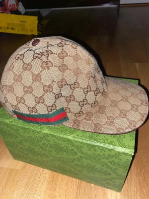 Gucci beige keps - Säljer en beige keps från Gucci i GG Supreme-mönster med klassiska gröna och röda ränder på sidan. Kepsen har böjd skärm och justerbart band bak i läder. Materialet är canvas med läderdetaljer och kepsen har en riktigt lyxig vibe.