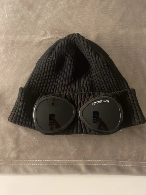 Svart Goggle-mössa från C.P. Company - Säljer en svart ribbstickad mössa från C.P. Company med de ikoniska goggle-linserna framtill. Mössan har uppvikt kant och är tillverkad i mjukt, stretchigt material som sitter skönt på huvudet. Perfekt accessoar för en streetwear-look.