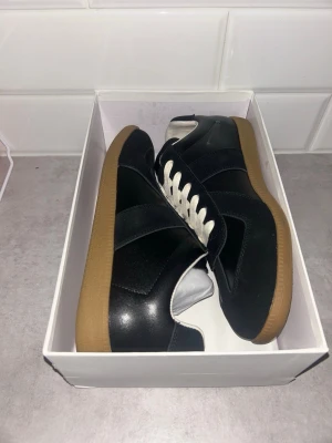 Svarta Maison Margiela - Snygga svarta sneakers med vit snörning och klassisk gummisula. Ovandelen är i en mix av mocka och läder för en stilren look. Perfekta för dig som gillar minimalistisk och tidlös design.