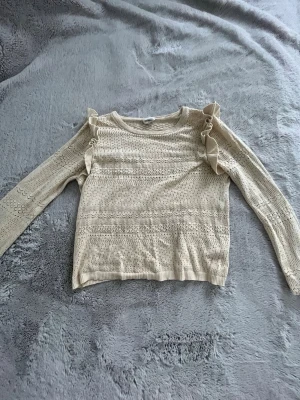 Beige stickad tröja med volangdetaljer - Söt beige stickad tröja med lång ärm och volanger längs axlarna. Tröjan har ett genombrutet mönster och ribbade muddar vid ärmslut och nederkant. Perfekt för dig som gillar romantiska detaljer och en mjuk, feminin stil.