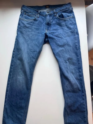 Blå jeans från Lee med rak passform - Säljer ett par klassiska blå jeans från Lee med rak passform och fem fickor. Jeansen har en snygg tvättad look och är tillverkade i slitstarkt denimtyg. Perfekta för dig som gillar en tidlös och enkel stil.
