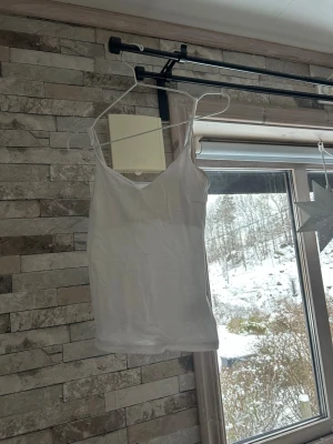 Vit linne med smala axelband - Enkel och stilren vit linne med smala axelband och v-ringad hals. Perfekt att styla lager på lager eller bära som den är under varmare dagar. Tidlös design som passar till många olika looks.