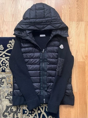 Moncler cardiganjacka i storlek S - Helt Ny - Denna Moncler cardiganjacka i storlek S kombinerar funktion och stil. Den har en quiltad design med en huva för extra värme och komfort. Perfekt för kalla dagar, och med sin svarta färg passar den både till vardags och mer avslappnade tillfällen. Jackan har också en praktisk armficka och en trendig siluett som gör den till ett utmärkt val för både stilmedvetna och praktiska användare.