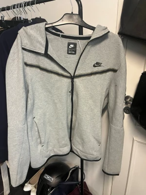 Grå hoodie med dragkedja från Nike - Snygg grå hoodie från Nike med svart logga på bröstet och hel dragkedja framtill. Hoodien har svarta detaljer längs dragkedjan och en klassisk huva. Perfekt för en sportig och avslappnad stil. Tillverkad i mjukt material för extra komfort.Helt okej skick, lite slitningar på texten och en liten fläck på armen som säkert går bort i tvätten