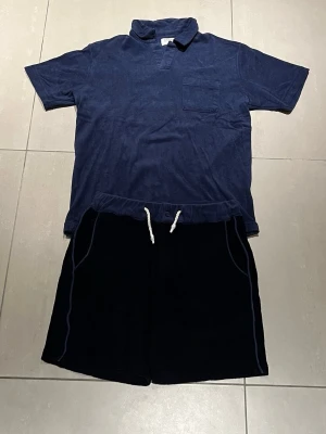 Mörkblå frotté pikétröja och shorts från Gabba - Mörkblå frotté pikétröja & shorts från Gabba med vit snörning i midjan och diskreta sömdetaljer längs sidorna. Sköna och avslappnade, perfekta för sommaren. Tillverkade i mjukt material som känns bekvämt mot huden.