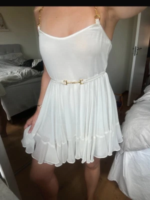 Vit kortklänning med gulddetaljer - Vit klänning från Ida Sjöstedt med volanger och gulddetaljer. Perfekt som studentklänning. Har inbyggd underkjol så är ej genomskinlig. Knyte i midjan. 