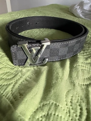 Svart rutigt skärp i skinn - Snyggt svart och grått skärp med klassiskt rutigt mönster och stor logga i silver på spännet. Tillverkat i skinn och har exklusiv känsla, perfekt för att lyfta vilken outfit som helst. Skriv vid fler frågor och bilder. 