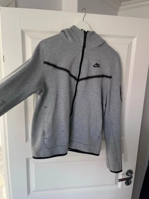Grå Nike hoodie med dragkedja - Snygg grå hoodie från Nike med svart dragkedja som går snett över bröstet och svarta detaljer. Hoodien har huva, två fickor framtill och en diskret Nike-logga på bröstet. Tillverkad i mjukt sweatshirtmaterial som är perfekt för chill eller träning.