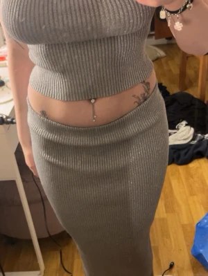 Glittrig silver långkjol med matchande topp  - Säljer en glittrig silverfärgad långkjol med ribbad struktur.  Med passande topp. Kjolen är figurnära och har en stretchig passform som framhäver siluetten. Perfekt för dig som vill sticka ut med en trendig och snygg look.