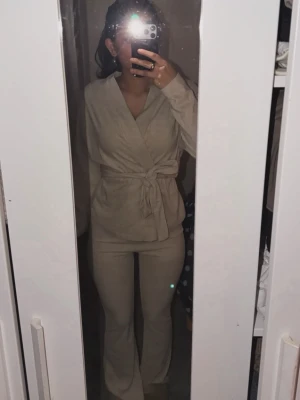 Beige set från Cider med omlottopp - Snyggt tvådelat set från Cider i beige färg. Setet består av en omlottopp med knytband i midjan och matchande byxor med utsvängda ben. Mjukt och stretchigt material som sitter bekvämt och ger en stilren look. Pluss en linne som är under koftan ingår. Prislapp med helt nya.