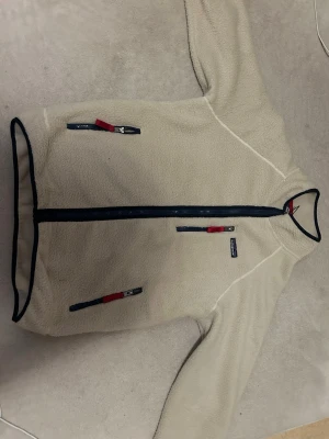 Beige fleecejacka från Patagonia - Säljer en beige fleecejacka från Patagonia med hel dragkedja framtill och kontrasterande mörkblå detaljer. Jackan har tre fickor med dragkedja, där dragarna har röda detaljer. Perfekt för lager-på-lager och outdoor-vibe.