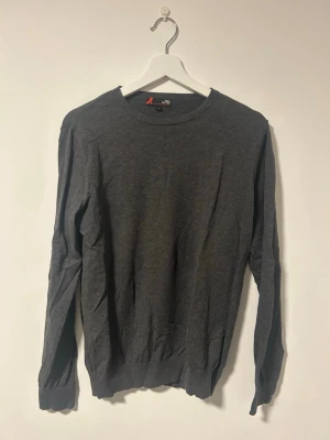 Mörkgrå stickad tröja från Dressmann - En stilren mörkgrå stickad tröja tillverkad i bomull med rund halsringning och långa ärmar. Tröjan har ribbade muddar vid ärmslut och nederkant. Tröjan har mest legat i garderoben och är även väldigt elastisk. Pris kan diskuteras. Fler bilder skickas vid behov.