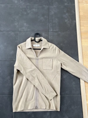 Beige overshirt från Samsøe Samsøe - Stilren beige overshirt från Samsøe Samsøe. Modellen har krage, bröstficka och hel dragkedja framtill. Perfekt att bära öppen eller stängd för en avslappnad look. Använd väldigt få tillfällen.