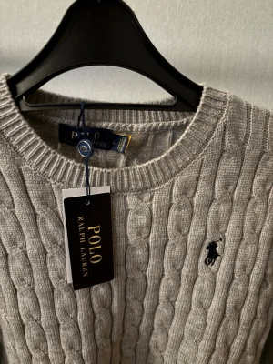 Grå kabelstickad tröja Polo Ralph Lauren - Kabelstickad grå tröja från Polo Ralph Lauren med rund halsringning och broderad svart logotyp på bröstet. Tröjan har ribbade muddar och klassisk passform, perfekt för en stilren look.