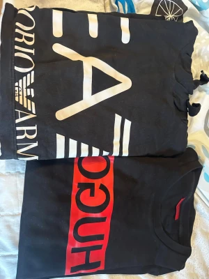 Svarta t-shirts från Hugo Boss & Armani - Två svarta t-shirts, en från Hugo Boss med röd logga framtill och en från Armani med stor vit logga och grafiskt tryck. Båda har klassisk rund hals och korta ärmar. Perfekta för dig som gillar stilrena märkesplagg med tydliga loggor.