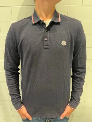 Mörkblå långärmad piké från Moncler - Snygg mörkblå långärmad pikétröja från Moncler. Den är storlek M men mocker sitter lite mindre i strl han på bilden är 181 och väger 70 kg. Bara höra av er vid minsta fundering 