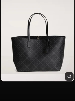 Svart shoppingväska från By Malene Birger - Stilren svart shoppingväska från By Malene Birger med diskret rutmönster och två långa handtag. Väskan är rymlig och har en klassisk, fyrkantig form. Tillverkad i slitstarkt konstläder och har en dekorativ tag med logga framtill.