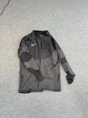 Svart träningsjacka från Nike - Snygg svart träningsjacka från Nike med hel dragkedja och diskret logga på bröstet. Jackan har långa ärmar med paneler i olika nyanser av svart och grått, samt praktiska sidofickor. Perfekt för träning eller uppvärmning.