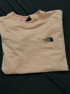 Ljusrosa t-shirt från The North Face - Snygg ljusrosa t-shirt från The North Face med klassisk logga tryckt på bröstet. T-shirten har rund halsringning och är tillverkad i mjuk bomull för en skön känsla. Perfekt till jeans eller shorts för en clean och enkel look.