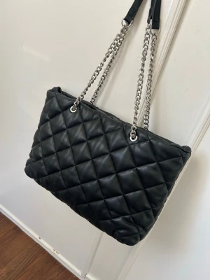 Svart quiltad handväska med kedjerem - Stilren svart handväska i quiltat konstläder med silverfärgade kedjeremmar. Väskan har en mjuk, puffig look och rejäla axelband i metall och läderdetaljer. Perfekt för dig som gillar en trendig och klassisk accessoar.