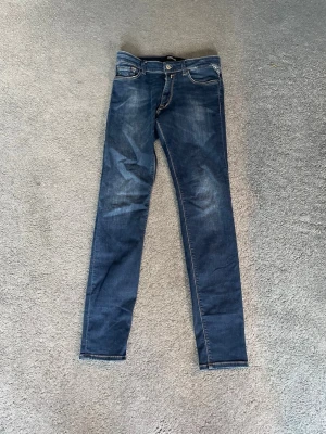 Mörkblå skinny jeans från Replay - Snygga mörkblå skinny jeans från Replay med klassisk femficksmodell och slitna detaljer på låren. Jeansen har vita kontrastsömmar och metallknapp i midjan. Perfekta för dig som gillar en smal passform och tidlös stil.