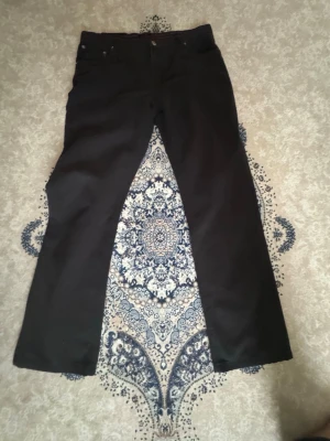 Svarta bootcut jeans från River Jeans - Svarta jeans från River Jeans med bootcut-passform och klassisk femficksdesign. Snygga röda detaljer på insidan av linningen. Perfekta för dig som gillar en stilren look med lite extra edge.