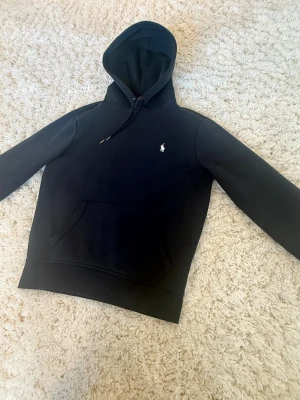 Svart hoodie från Polo Ralph Lauren - Mörkblå hoodie från Polo Ralph Lauren med klassisk vit broderad logga på bröstet. Hoodien har dragsko i huvan och en stor känguruficka framtill. Tillverkad i mjukt bomullsmaterial för en skön och avslappnad stil.