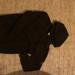 Svart hoodie från Stone Island - Säljer en svart hoodie från Stone Island med klassisk huva och dragsko. Tröjan har lång ärm och den ikoniska Stone Island-loggan på vänster ärm. Perfekt för dig som gillar streetwear och enkel stil.