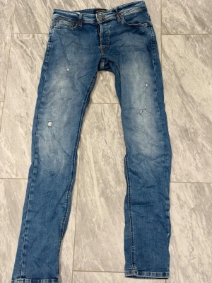 Blå slitna slim fit jeans - Snygga blå slim fit jeans med slitningar och lätt tvättad look. Jeansen har klassisk femficksdesign, låg midja och detaljerade slitningar framtill för en trendig vibe. Perfekta för en avslappnad streetstyle.