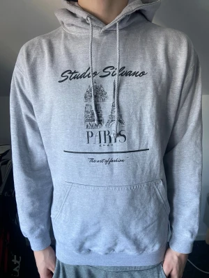 Grå hoodie Studio Silvano Paris - Snygg grå hoodie från Studio Silvano med tryck av Eiffeltornet och texten 'Paris' på bröstet. Hoodien har huva med dragsko och en stor magficka. Tillverkad i mjukt material, perfekt för en avslappnad stil. Storlek M, passar dig som är runt 180, köpt för 500 säljer för 329.