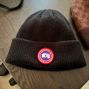 Svart  mössa Canada Goose båda 600  - Svart ribbstickad mössa från Canada Goose med klassisk rund logotyp-patch framtill. Tjockt uppvik och stilren design som passar perfekt till kalla dagar. Materialet ser ut att vara ull eller ullblandning för extra värme. Testade bara . 300 st