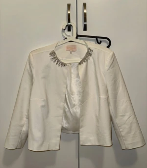 Vit kavaj med pärlor från Mohito - Snygg vit kavaj från Mohito med glittrande pärldetaljer runt halsringningen. Kavajen har lång ärm, öppen framtill och ett lyxigt satinfoder med diskreta prickar. Perfekt för dig som vill sticka ut med stilrena detaljer.