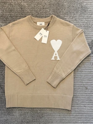 Beige stickad tröja från Ami - Snygg beige stickad tröja från Ami med rund halsringning och ribbade muddar. På bröstet finns ett vitt hjärta och bokstaven A som ger en unik touch. Perfekt för dig som gillar stilrena plagg med en twist.