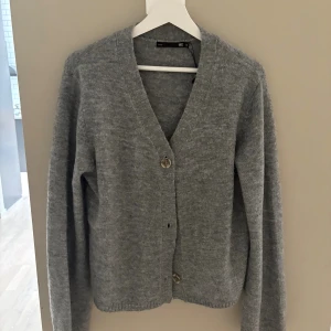 Grå stickad kofta från 157 - Mysig grå stickad kofta från 157 med v-ringning och 3 knappar framtill. Koftan har lång ärm och en klassisk, enkel design som passar perfekt till lager-på-lager. Materialet är mjukt och ger en chill vibe till din outfit.