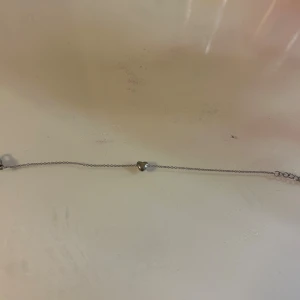 Silverfärgat armband med hjärta - Ett tunt och stilrent armband i silverfärg med ett litet hjärta i mitten. Justerbar kedja med en rund bricka i änden som har en ingraverad bokstav. Perfekt för dig som gillar enkla och söta smycken.