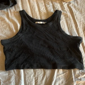 Svart ribbad croppad linnetopp lager 157 - Säljer en svart croppad linne från 157 i ribbad bomull. Toppen är ärmlös med breda axelband och har en tight passform som sitter snyggt. 
