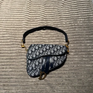 Fin väska  -  blå canvas med monogrammönster och detaljer i mörkblått skinn. Väskan har guldiga metalldetaljer, inklusive det klassiska D-spännet. Snygg och unik form med bred axelrem och extra avtagbar rem.