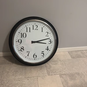 Stor rund väggklocka med svart ram - Säljer en stor och stilren rund väggklocka med svart ram och vit urtavla. Klockan har tydliga svarta siffror och visare, perfekt för att hålla koll på tiden i köket, vardagsrummet eller pluggrummet.