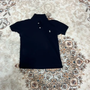 Mörkblå pikétröja från Ralph Lauren - Klassisk svart pikétröja från Ralph Lauren med vit broderad logga på bröstet. Tröjan har korta ärmar, krage och två knappar framtill. Perfekt för en clean och stilren look.