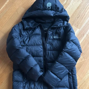  Sail Racing JR Cloud Down Hood - Svart - Svart dunjacka med luva från Sail Racing. Jackans vaddering består av 90% dun och 10% fjäder. Framtill finns fickor med dragkedja och märkets logga är tryckt och placerad på bröstet. Jackan andas och är vindtät. Vill man ha en varm och stilren jacka så är denna perfekt.  Jackan har en liten lagningslapp, se bild 5  https://kidsbrandstore.se/products/jr-cloud-down-hood-27326